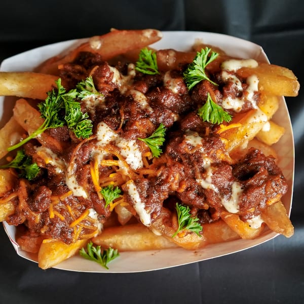 Chili cheese fries topped with white sauce and fresh parsley in a white paper tray.