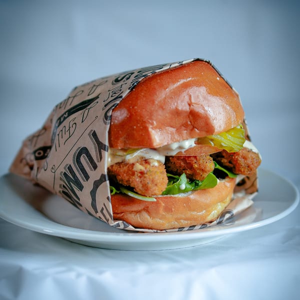 A fried chicken sandwich with pickles, lettuce, and sauce, wrapped in paper on a white plate.