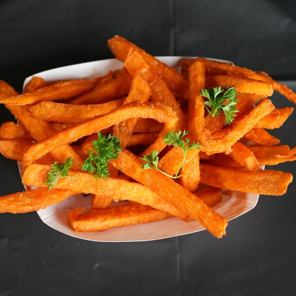 Crispy sweet potato fries garnished with fresh parsley in a white paper tray on a dark background.