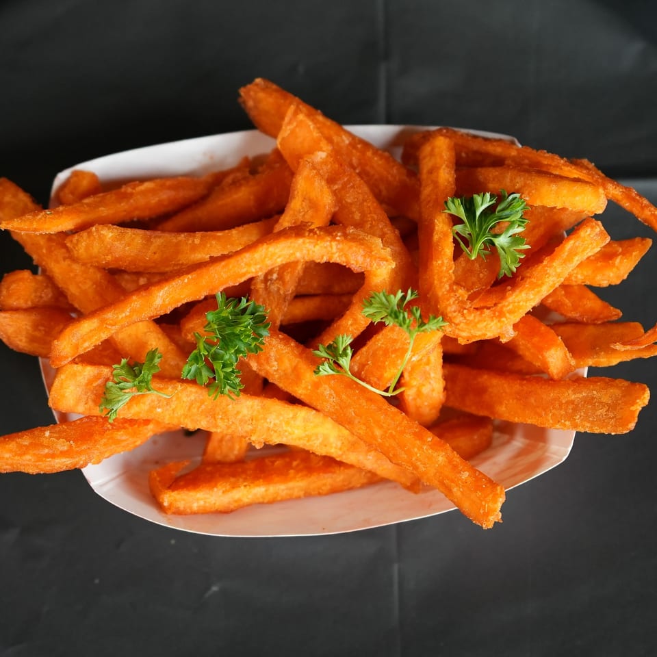 Crispy sweet potato fries garnished with fresh parsley in a white paper tray on a dark background.