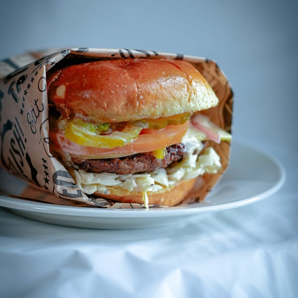 A juicy burger with pickles, tomato, and sauce, wrapped in paper, sits on a white plate.