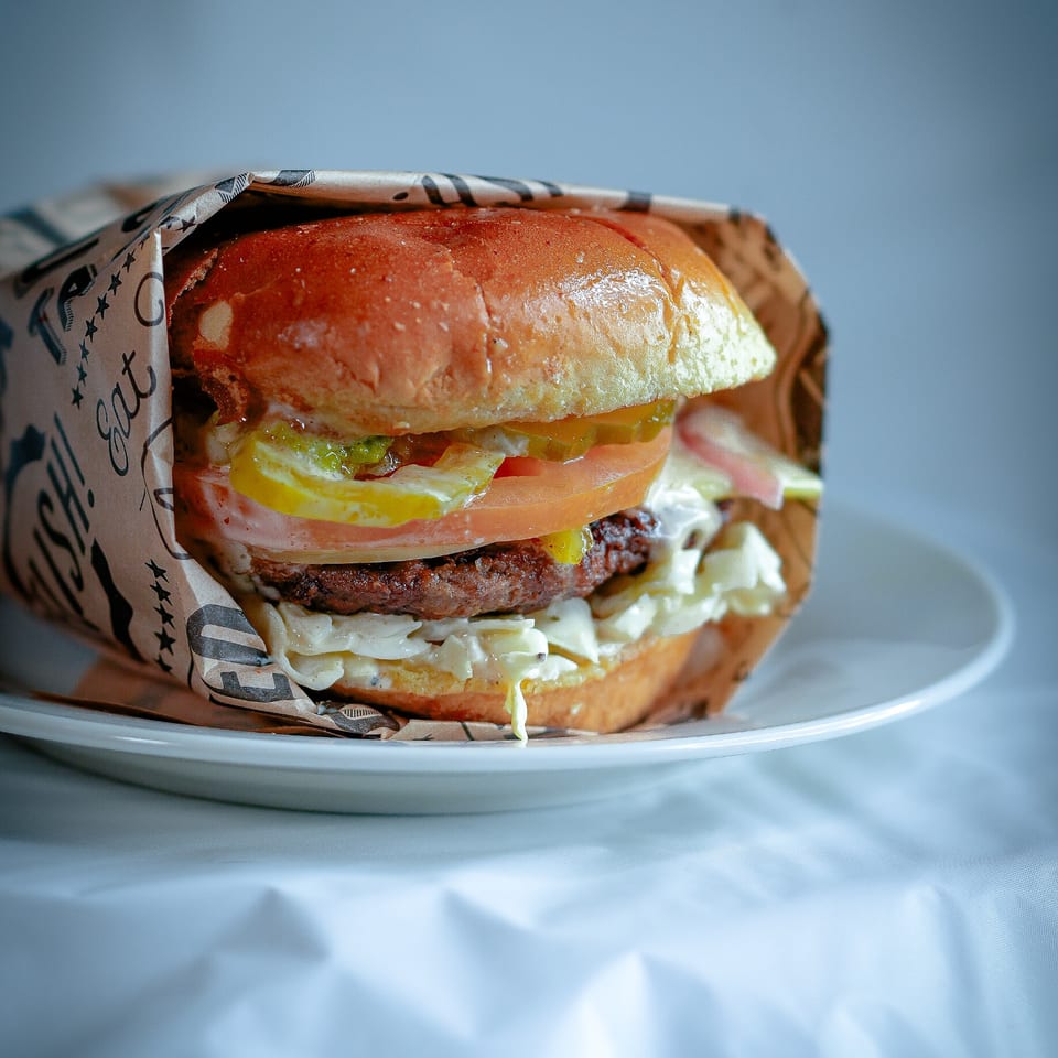 A juicy burger with pickles, tomato, and sauce, wrapped in paper, sits on a white plate.