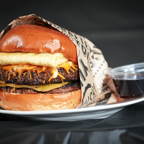 A double-patty burger with cheese, egg, and sauce, wrapped in paper, on a white plate.